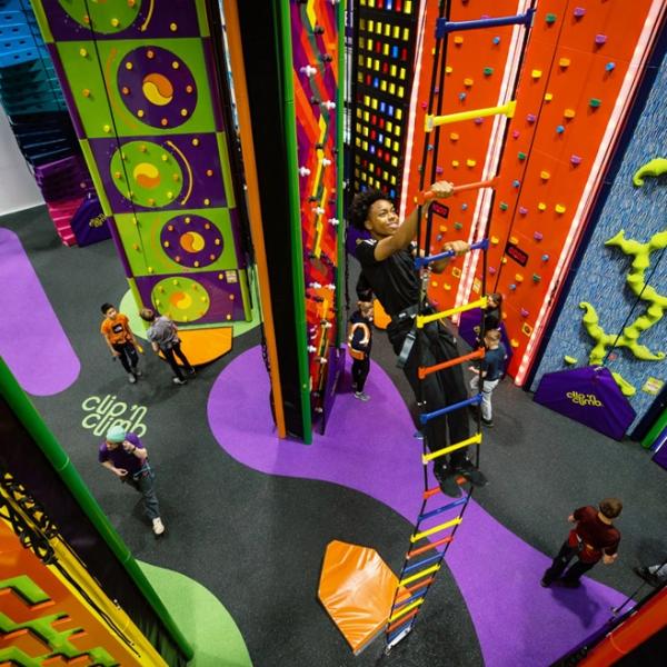 A colourful indoor climbing centre with various themed climbing walls, a hanging ladder, and several people climbing or waiting on the padded floor below.