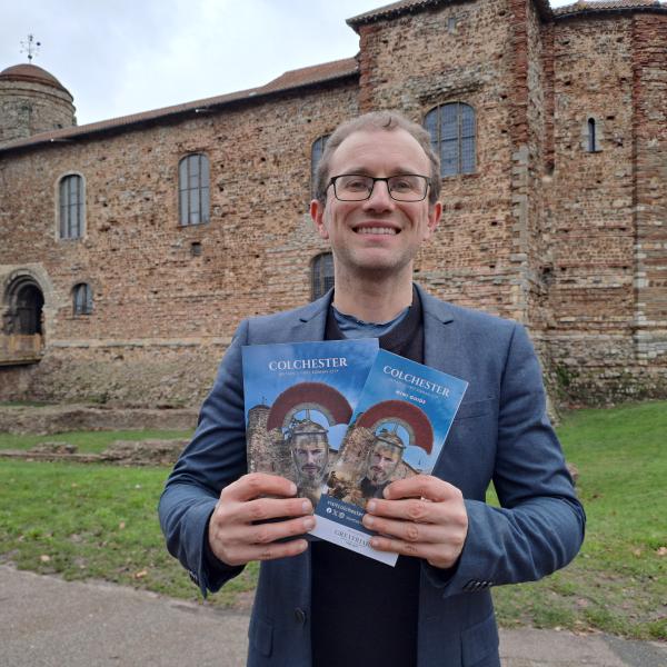 Cllr Lee Scordis holding Visit Colchester Guide in front of Colchester Castle