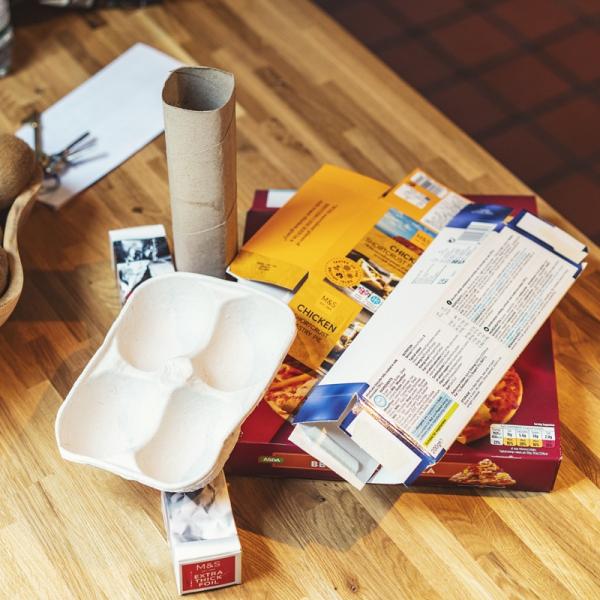 Recyclable cardboard and paper items laid out on a wooden kitchen counter beside a bowl of fruit.