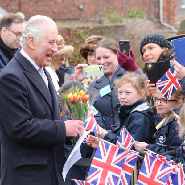King Charles III holds a bouquet of yellow and red tulips while greeting a line of schoolchildren holding small Union Jack flags, with adults gathered behind them taking photos during an outdoor public visit.