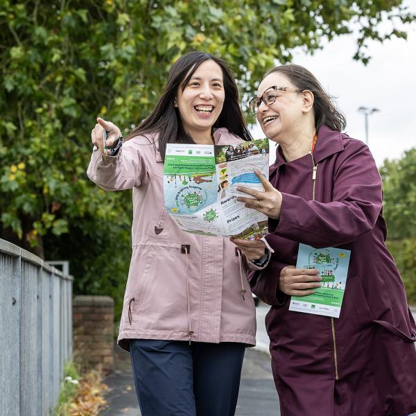 Two people standing outdoors looking at a printed map or leaflet. One person is pointing ahead while the other holds the leaflet open as they discuss the route. They are on a pavement next to a railing with trees and greenery in the background.