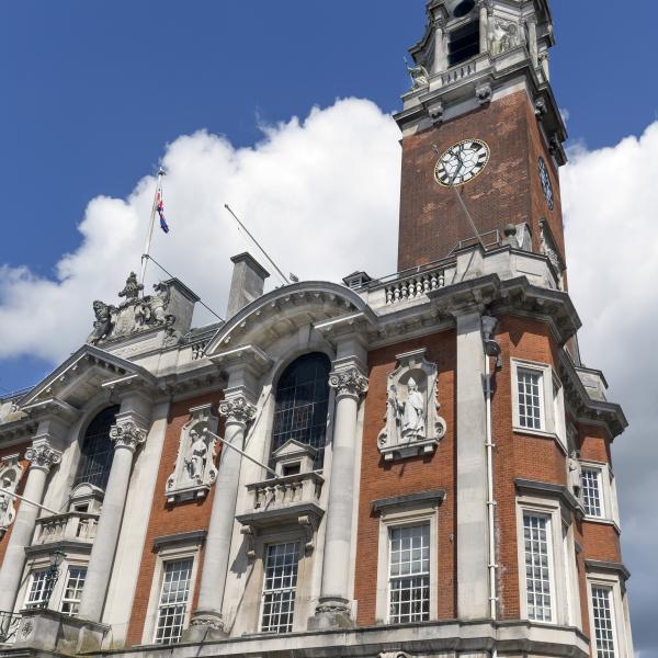 A tall historic brick building (Colchester Town Hall) with ornate stone detailing, arched windows, and classical statues set into the façade. A prominent clock tower rises above the structure, topped with decorative architectural features. The sky behind it is bright blue with scattered white clouds.
