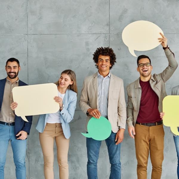 Five people standing against a gray textured wall, each holding large colorful speech bubble cutouts in various shapes. The speech bubbles are in shades of blue, yellow, green, and beige, suggesting themes of communication, conversation, or collaboration. The individuals are dressed in business-casual attire, and the overall setting conveys a modern, professional environment.