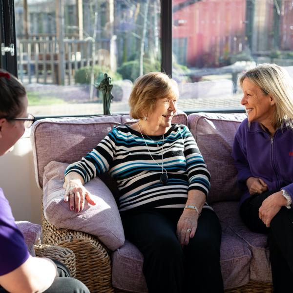 Two Helpline staff members talking to a Helpline customer at their home. Sitting on a sofa and chair in a living room setting. There is a large window behind them showing a street scene.