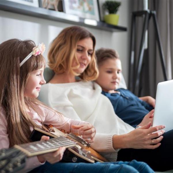 A scene of three individuals sitting together on a sofa in a living room. One person holds a laptop, while another sits nearby with a guitar resting across their lap. The room features soft natural light, wall décor on a shelf, and a floor lamp in the background.