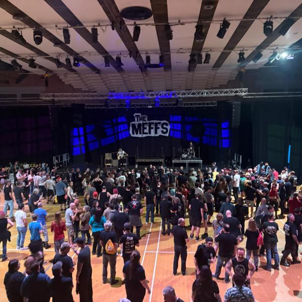 A large crowd stands inside Charter Hall facing a stage with “The Meffs” backdrop.