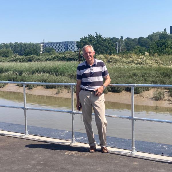 Cllr Sunnucks standing on a paved quayside path by a metal railing beside a tidal river, with marshland, trees and modern buildings in the background under a clear blue sky.