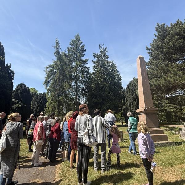 A group of people gathered around a tall obelisk monument among trees in Colchester Cemetery.