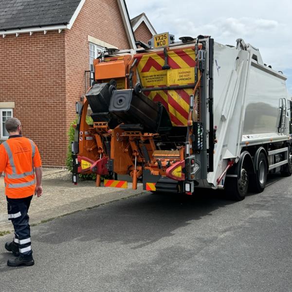 Refuse collection lorry collecting bins outside houses, with a worker in high-visibility clothing nearby.