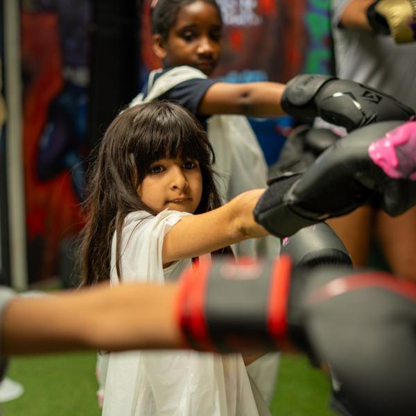 A group of children wearing boxing gloves practise punches on pads in a gym.