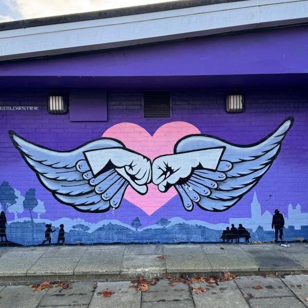 A large mural painted on a purple brick wall showing two white winged hands touching knuckles in front of a pink heart. Silhouettes of people, trees and a city skyline run along the bottom of the artwork, with a paved walkway in the foreground and a cloudy sky above.