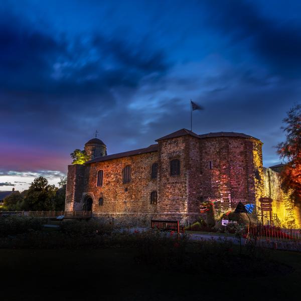 Colchester Castle illuminated at dusk beneath a dramatic blue and purple evening sky. The historic stone castle is lit with warm amber, pink and gold floodlights that highlight its textured walls and arched doorway. Trees frame the scene on both sides, with one brightly illuminated in orange and red light. A flag flies from the castle roof, and gardens, pathways, benches and visitor signage are visible in the foreground. 