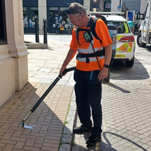 A person uses a long tool to remove chewing gum from a pavement beside a street.