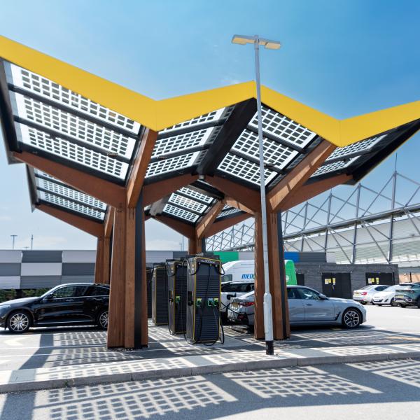 Solar-panel canopy sheltering electric vehicle charging points in a car park beside a large building.