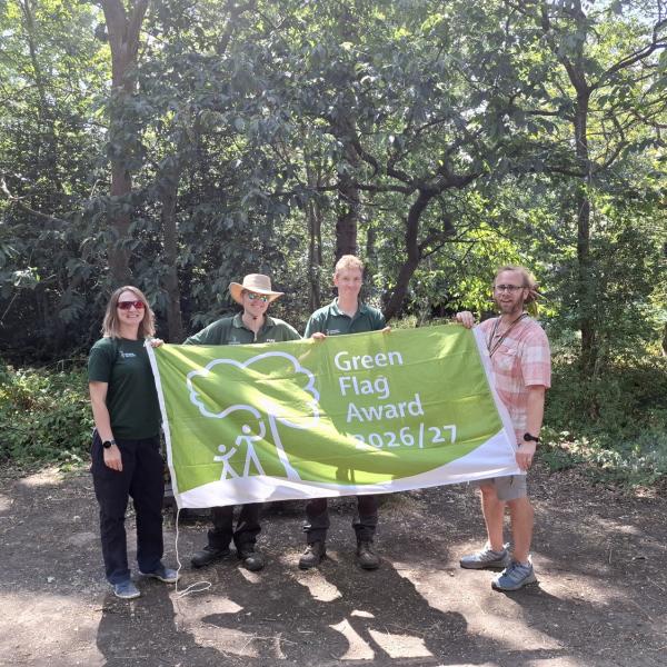 Four people hold a Green Flag Award 2026/27 banner in a wooded park.