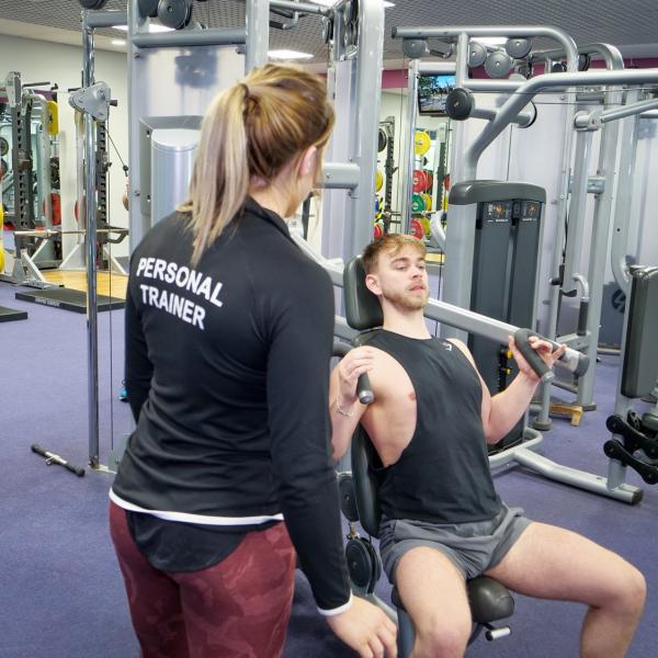 Person in 'Personal Trainer' top observing another using a chest press machine