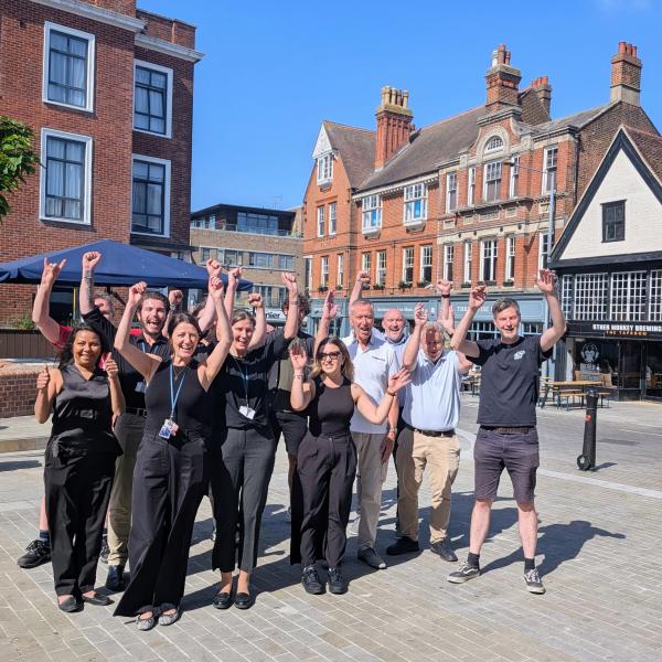 A group of around 12 people stand together in a paved town square with both arms raised in the air. The group is positioned in front of a row of historic red-brick and timber-fronted buildings under a clear blue sky. Outdoor café umbrellas and a small tree are visible on the left, while a building with signs reading “Other Monkey Brewing” appears in the background on the right. The scene is brightly lit by sunshine, with long shadows cast across the square.
