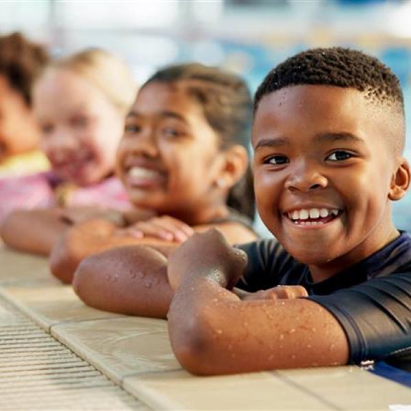 A group of children leaning on the edge of an indoor swimming pool in a line.