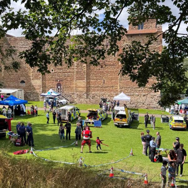 A community event on a grassy lawn beside a large historic stone building, with display tents, emergency service vehicles and groups of people.