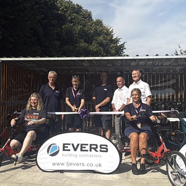 A group of people at a ribbon-cutting ceremony beside adapted cycles in a covered outdoor area.