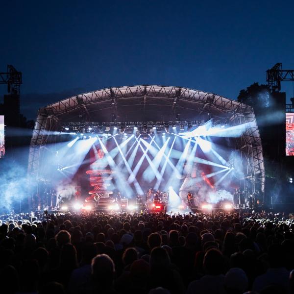 A large outdoor concert stage illuminated by bright lights, with a crowd gathered in front at night.
