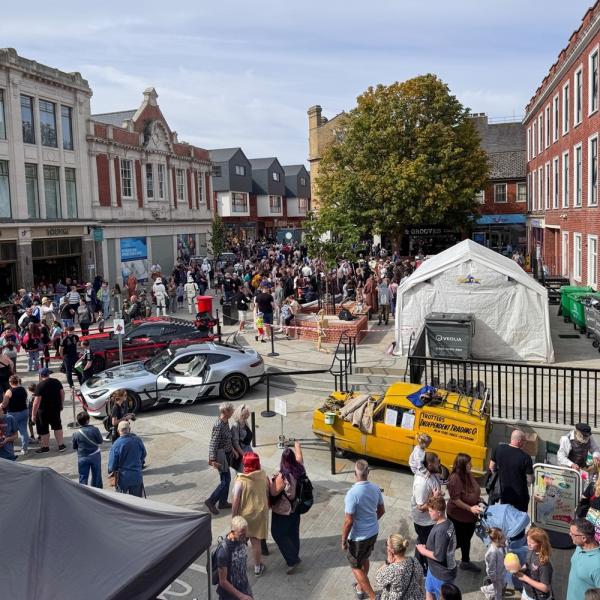 A busy city centre square with crowds, display vehicles, market stalls and surrounding buildings.