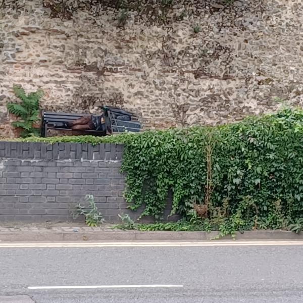 A person sleeping on a bench beneath Colchester's Roman Wall