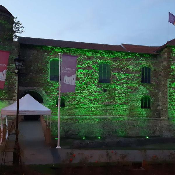 Colchester Castle at dusk, illuminated with green lighting across its historic stone walls. A pathway and bridge lead to the main entrance beneath a white canopy. Two flags reading "Colchester Castle" stand in front of the building, and a rainbow flag flies from a flagpole on the roof. Trees frame the castle on both sides beneath an overcast evening sky.