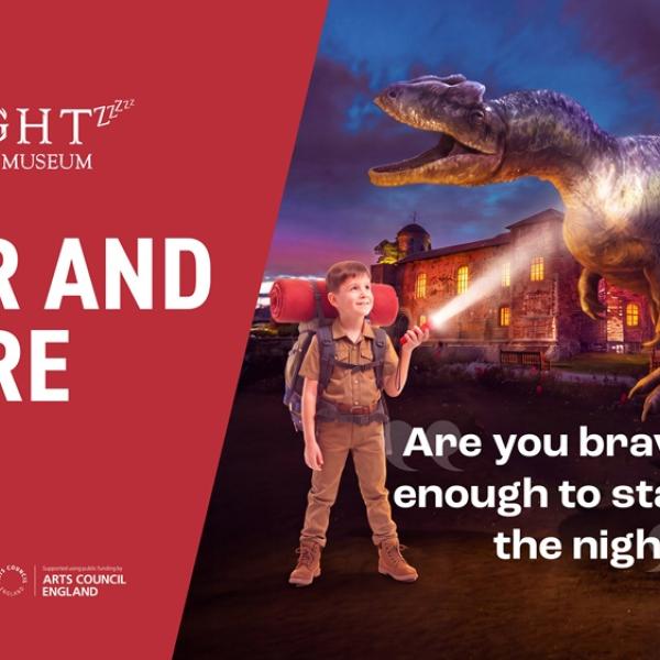 Two people with backpacks and torches stand in front of a dinosaur and museum at night.