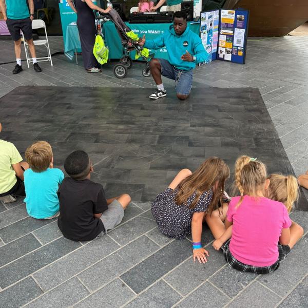 A person kneels on a mat demonstrating moves to seated children at an event.