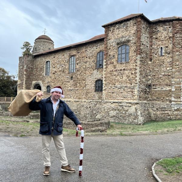 Cllr Darius Laws wearing a Christmas hat and carrying a sack of presents in front of Colchester Castle